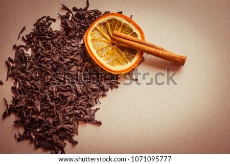 Black, dry leaves of tea, next to a circle of orange and a stick of cinnamon. A fragrant choice tea, a traditional drink. Light background, warm toning, vignetting, copy space.