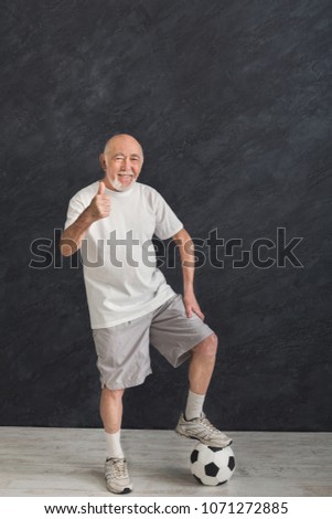 Smiling senior man posing with soccer ball showing thumb up gesture, black background. Hobby, active lifestyle in any age and motivation, copy space