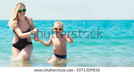 Beautiful young mother and small son holding hands running on the waves on the beach.Fun,family,friendly summer vacation.