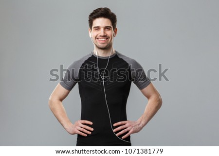 Photo of happy young sportsman standing isolated over grey wall background looking camera listening music with earphones.