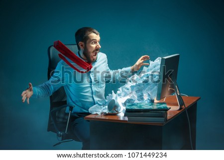Young stressed handsome businessman working at desk in modern office shouting at laptop screen and being angry about e-mail spam. Collage with a mountain of crumpled paper. Business, internet concept Royalty-Free Stock Photo #1071449234