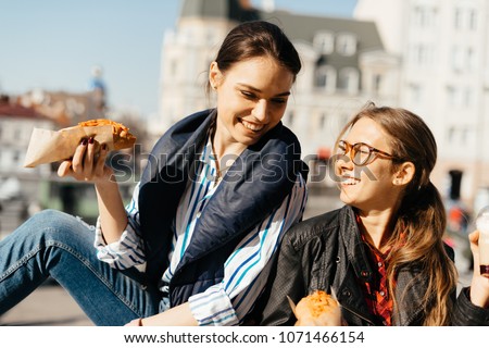 Dark hair woman and blonde woman models with hot-dogs and coffee to go in the city park smiling