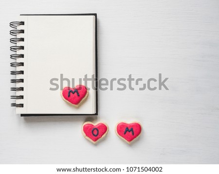 Fresh delicious cookies in the shape of a heart with the word MOM, covered with glaze, an album with a blank page for Your inscription.White isolated background.Top view.Concept of a happy family