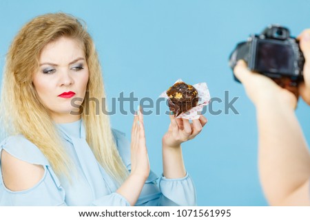 Woman refuses to eat chocolate cupcake, man taking picture of her. Blue background.