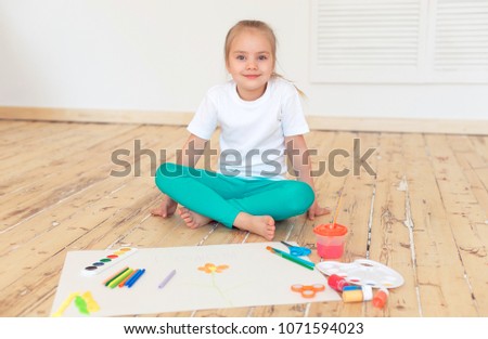 Little blonde girl paintson big white paper sitting on the floor indoors.