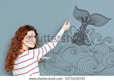 Beautiful painting. Creative talented artist looking glad while standing near the wall and painting a big beautiful whale on it