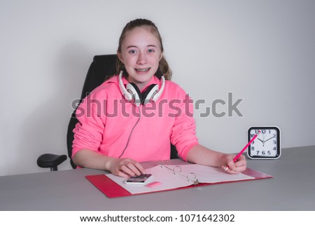 Happy teenage girl sitting at a desk studying/doing home work while holding a pencil with headphones around neck.
