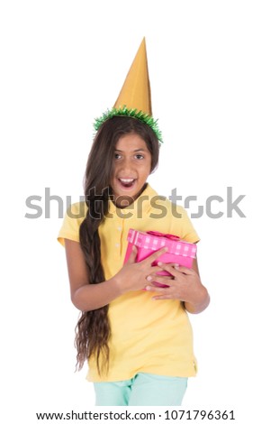 Little beautiful girl wearing birthday cone and holding gift box she received smiling with open mouth, isolated on a white background.