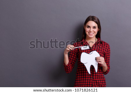 A young cheerful woman holds a paper image of a tooth and a toothbrush on a gray background. Healthcare concept.