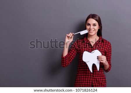 A young cheerful woman holds a paper image of a tooth and a toothbrush on a gray background. Healthcare concept.