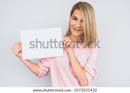 Closeup shot of beautiful happy smiling blonde female wearing pink sweater, posing with white blank paper with copy space for your advertisement information and looking at camera, against white wall. 