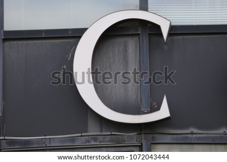 Close up view of the alphabet letter C, fixed on a building wall. Metallic element with curved shape. Modern design with grey, white and black colours. Abstract image of an uppercase character. 