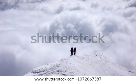 two female mountain climbers on a narrow summit ridge high above cloud banks in the valleys below Royalty-Free Stock Photo #1072067387