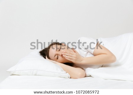 Calm young brunette woman lying in bed with white sheet, pillow, blanket on white background. Yawning beauty female spending time in room. Rest, relax, good mood concept. Copy space for advertisement