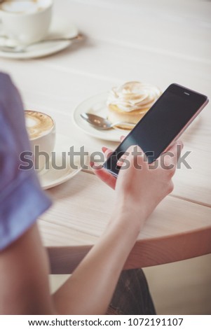 Hand with a phone, cup of cappuccino and a piece of dessert on the plate, soft focus background