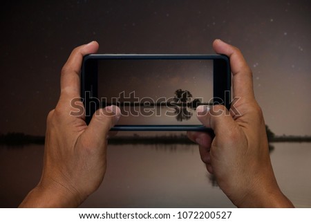 Man hand holding and using mobile,cell phone,smart phone photography and Lake in the night with the stars in the sky and the reflection of the trees and the sky.