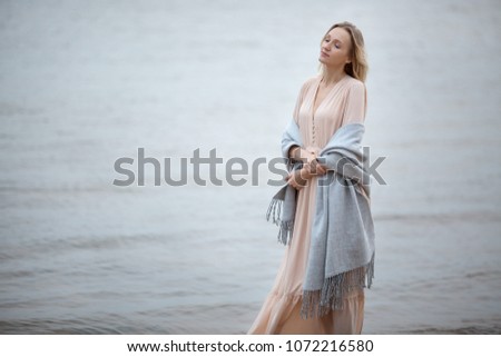 Young blond woman in a long pink dress is wrapped up in a warm blanket walking along the seaside enjoying nature.