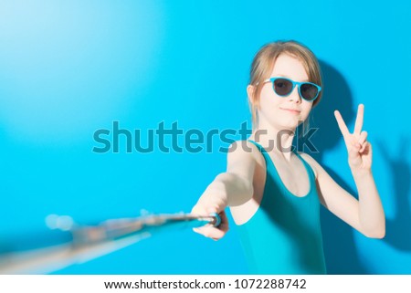 Adorable girl in swimsuit with sunglasses using selfie stick and taking selfie showing two fingers.