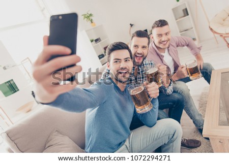 Stylish, attractive, modern positive guys sitting in living room, having mugs with lager in hands, shooting selfie on smart phone, shouting, screaming, enjoying sport football competition