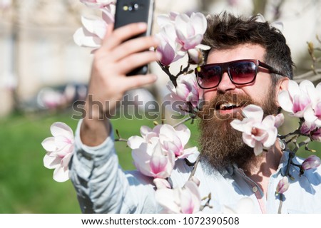Man with beard and mustache wears sunglasses on sunny day, magnolia flowers on background. Blogger concept. Hipster happy in stylish sunglasses, taking selfie photo, streaming video on smartphone.
