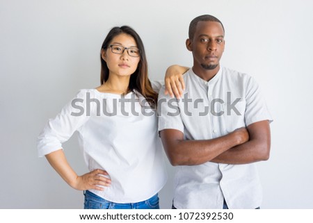 Serious confident young multiethnic couple looking at camera and standing in studio. Modern Asian woman leaning on boyfriends shoulder, Interracial friendship concept