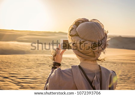 Young woman wearing turban (back view) taking picture of sunset in desert, Siwa, Egypt