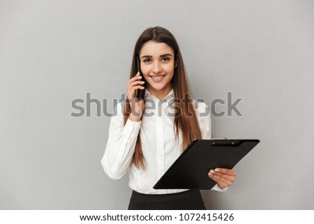 Photo of businesslike happy woman in white shirt and black skirt holding clipboard with files in office and calling with mobile phone isolated over gray background