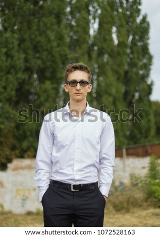 Young guy in white shirt and black trousers. Handsome smart man in glasses standing outside with green trees on background. Business portrait. Vertical photo.