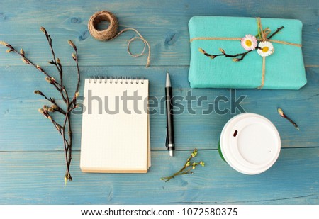 Copy book with empty shirt, cup of coffee, gift box wrapped with blue cloth, branch of spring tree with cute buds, dry flower of pink rose. Objects on wooden background, overhead view