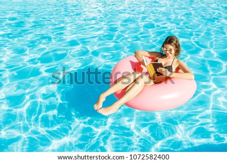 Beautiful happy woman reading a book with inflatable ring relaxing in blue swimming pool.
