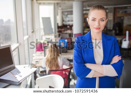 A portrait of business lady standing neat window with her hands crossed. She is looking straight to the camera. Girl looks confident and very good.