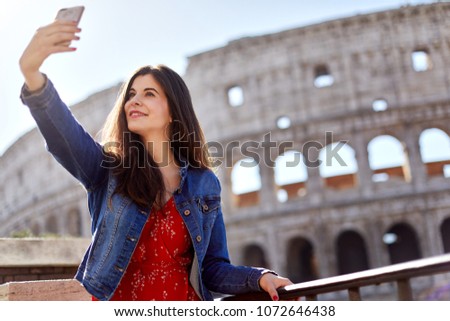 Brunette in dress and denim using phone and taking selfie on background of Colosseum in sunlight.