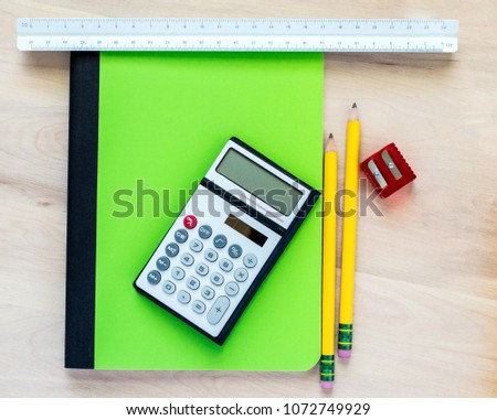 2 yellow pencils, a red pencil sharpener, a triangular ruler, a small calculator and a green notebook on a wooden desk