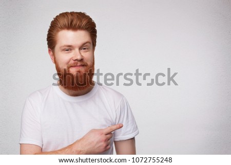 Indoor shot of young excited surprised redhead guy with beard point with a finger aside, smiles and feels happy