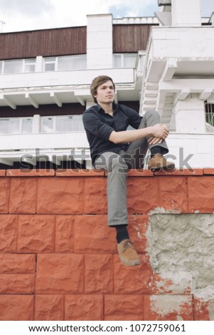 young brunette man with a disheveled hairstyle posing near an old brick building on a summer day. street style in clothes: office pants and a wide shirt. emotional portrait of a student. model test