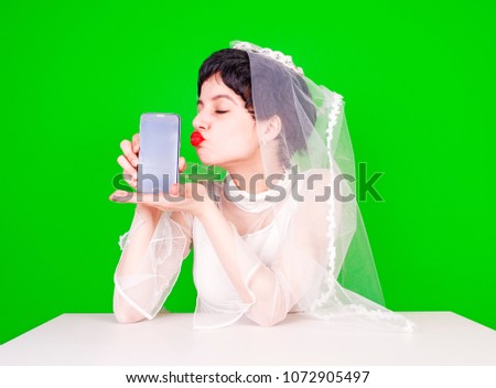 Stunning bride shows on the hand the screen of the phone and kisses it sitting at the table on a green background. Good news. Positive concept