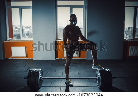 african american athletic man in sport mask put his foot on barbell waiting and preparing before lifting heavy barbell. deadlift with barbell in mask. bent over the barbell. opposite window