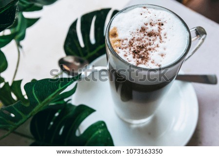 A cup of Hot cappuccino latte coffee with cinnamon sprinkles on top on wooden white dinner table with a green leaf in a  background