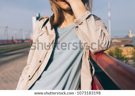 Candid lifestyle cropped close-up photo of young woman standing on the bridge smiles and covers her face from sunlight at sunny spring day.