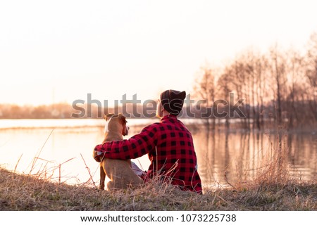 Young man and pet dog sit by lake at sunset in spring or autumn. Male person and staffordshire terrier puppy enjoy golden hour at dawn of a bright sunny day near river Royalty-Free Stock Photo #1073225738