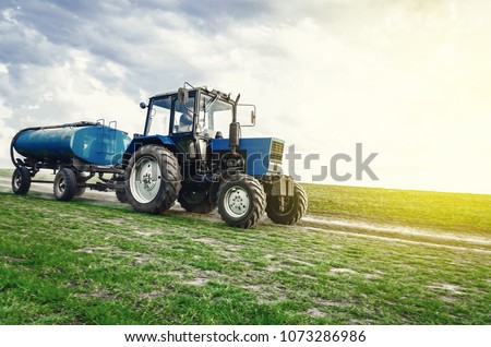 tractor of blue color with a barrel trailer rides along the spring field along the road. Successful farming.
