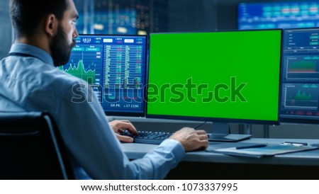 Over the Shoulder View of Stock Market Trader Working on a Computer with Isolated Mock-up Green Screen and Second Display Showing Number Ticker with Graphs. In the Background Monitors with Information