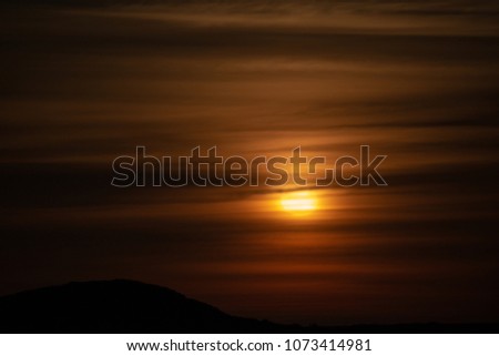 A background of a bright orange sunset or sunrise through thick heavy clouds over a silhouette of a landmass.  The clouds are horizontal lines with different shades of orange, brown, and black. 