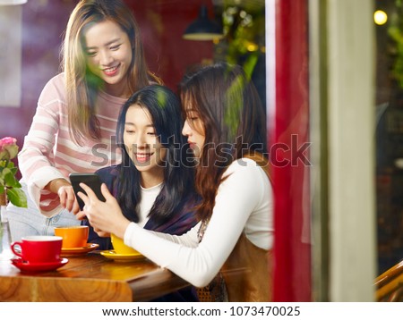 three happy beautiful young asian women sitting at table chatting talking playing with cellphone in coffee shop or tea house, shot through window glass.