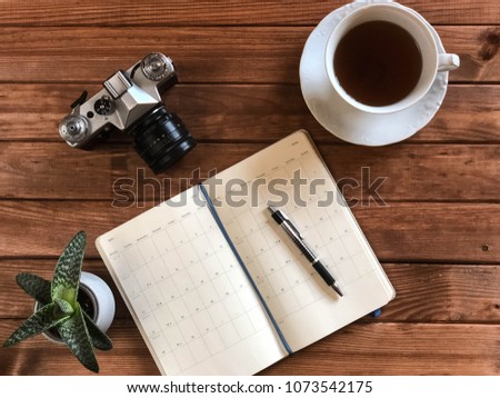 Aerial view on wooden working desk with open planner, retro camera, pencil, green succulent plant in a pot and porcelain white cup with black coffee. Concept of hipster workplace of a photographer