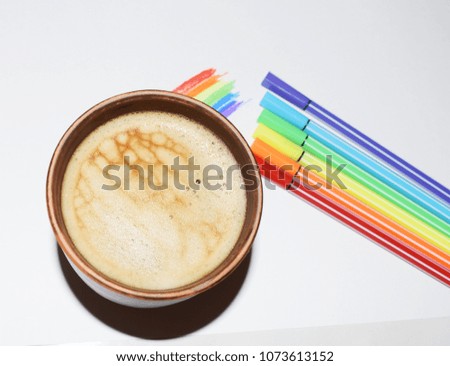Photo of a cup of coffee on a background of a painted rainbow and markers. A saturated coffee color with an airy foam and a bright aroma.
