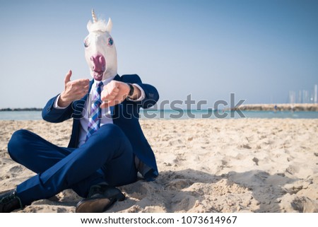 Stylish manager in comical mask and elegant suit sits on beach and looks on the watch. Funny man active gesticulating on background of sea and sky