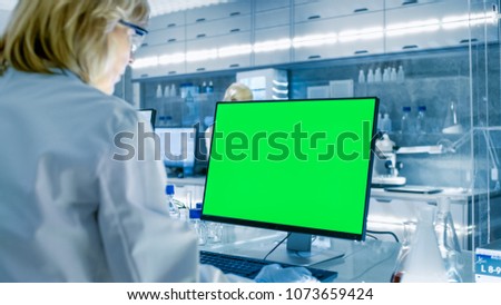 Female Scientist Working on her Computers (Mock-up Green Screen) In Modern Laboratory. Various Shelves with Beakers, Chemicals and Different Technical Equipment is Visible.