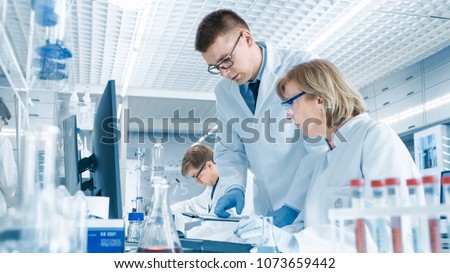 In Modern Laboratory Senior Female Scientist Has Discussion with Young Male Laboratory Assistant. He Shows Her Data Charts on a Clipboard, She Analyzes it and Enters It into Her Computer. Royalty-Free Stock Photo #1073659442