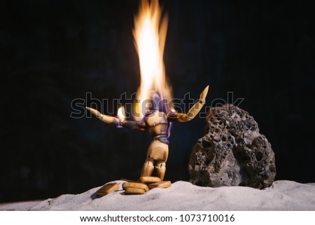 Conceptual picture of a burning wooden figure of a man standing on his knees with his hands up in front of a stone in a deserted landscape. The symbol of despair and appeals to the "higher forces"